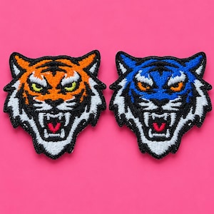 Op de afbeelding: Twee geborduurde patches in de vorm van tijgerkoppen. De ene patch toont een oranje tijger met zwarte strepen en gele ogen. De tweede patch toont een blauwe tijger met zwarte strepen en oranje ogen. Beide patches hebben witte en rode details.