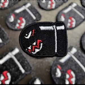 Op de afbeelding: Zwarte geborduurde patch in de vorm van een kogel met een witte rand en een rood en wit ontwerp erin.