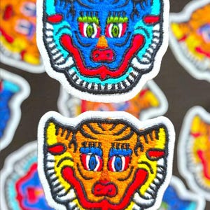 Op de afbeelding: Geborduurde patches met een gestileerd tijgergezicht. De patches hebben een witte rand en zijn in verschillende kleuren, waaronder blauw, oranje, rood en geel. De tijgergezichten hebben gedetailleerde kenmerken, waaronder ogen, neuzen en monden.