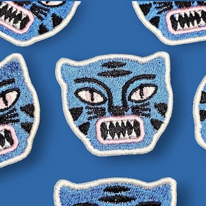 Op de afbeelding: Vijf blauwe geborduurde strijkpatches van een cartoontijger met zwarte strepen, witte tanden en roze tandvlees. De tijger heeft een woeste uitdrukking met grote, witte ogen.