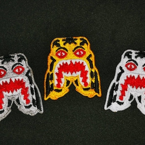 May include: Three embroidered patches featuring a cartoon shark with sharp teeth and a menacing expression. The patches are in black, white, and yellow.