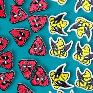 Op de afbeelding: Een verzameling rode en gele geborduurde strijk-op patches. De rode patches zijn driehoekig met een zwart-wit gezicht. De gele patches zijn in de vorm van een cartoonfiguur met een zwart gezicht en een gele hoed.