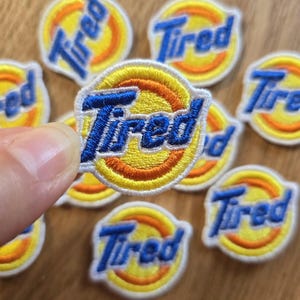 Może przedstawiać: Haftowane naszywki ze słowem "Tired" w niebieskim skrypcie. Naszywki mają żółty i pomarańczowy okrągły wzór z białym obramowaniem. Widocznych jest kilka naszywek, z których jedna jest trzymana w dłoni.