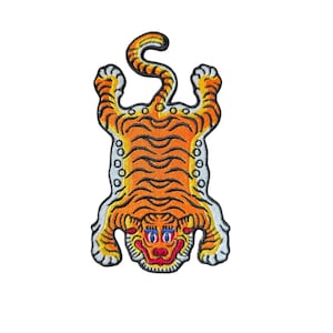 May include: Embroidered patch featuring a stylized tiger design. The tiger is depicted in orange and black with a white border. The tiger has a smiling face with blue eyes and a red mouth. The patch is likely for clothing or accessories.