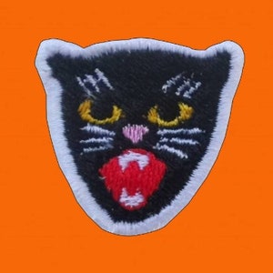 Op de afbeelding: Geborduurde patch met een zwarte kat met gele ogen, een roze neus en een rode mond met witte hoektanden. De kat is wit omlijnd en geplaatst tegen een oranje achtergrond. De patch heeft het woord "CAT" geborduurd.