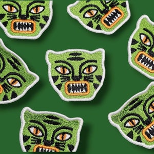 Op de afbeelding: Zeven groene en zwarte geborduurde strijkpatches met een cartoon-tijgergezicht met oranje tanden en witte ogen.