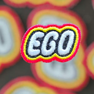 Op de afbeelding: Geborduurde patch met het woord "EGO" in wit, omlijnd met zwart, geel en roze. De patch heeft een afgeronde rechthoekige vorm. De achtergrond is wazig met vergelijkbare patches.
