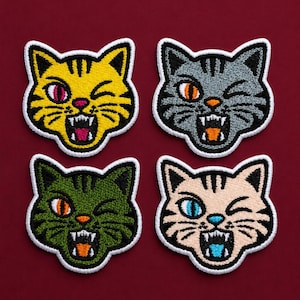 Op de afbeelding: Vier geborduurde kattenpatches met witte randen. Elke kat heeft een andere kleur: geel, grijs, groen en bruin. De katten hebben boze uitdrukkingen met open monden, tonen tanden en knipogen. De achtergrond is dieprood.