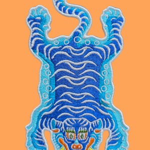 Könnte beinhalten: Gestickter Aufnäher in Form eines Tigers, mit Blau- und Weißtönen auf orangefarbenem Hintergrund. Das Tigerdesign umfasst detaillierte Gesichtszüge und Körpermarkierungen mit einem eingerollten Schwanz.