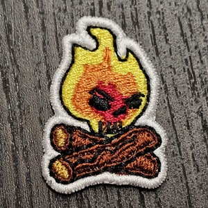 Op de afbeelding: Geborduurde strijkpatch met een schedel met boze ogen en een geel en oranje vlammenontwerp. De schedel zit op twee bruine boomstammen.