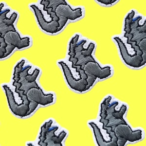 Op de afbeelding: Een herhalend patroon van grijze geborduurde patches van een Godzilla-achtig monster op een felgele achtergrond.