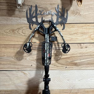 May include: A black and camouflage crossbow with a scope, mounted on a wooden wall. Above the crossbow is a metal cutout with the name "Luthy" and antler-shaped designs. Two animal skulls are mounted on the wall.