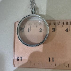 May include: A silver-tone, circular locket pendant with a clear glass front. The pendant, about 2.5 cm in diameter, hangs from a fine chain. The necklace is shown on a wooden ruler.
