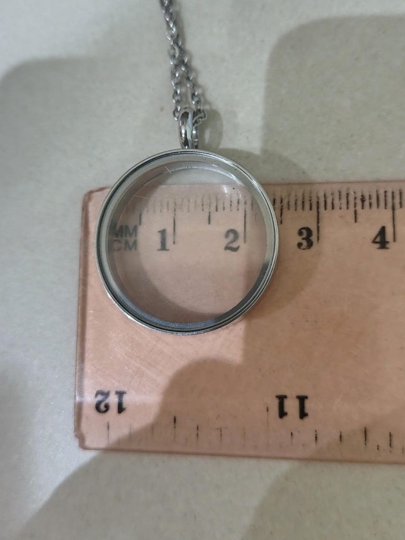 May include: A silver-tone, circular locket pendant with a clear glass front. The pendant, about 2.5 cm in diameter, hangs from a fine chain. The necklace is shown on a wooden ruler.