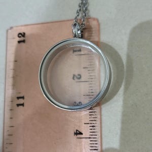 May include: A silver locket pendant with a clear glass centre, hanging from a fine chain. The locket is placed over a ruler, with measurements visible in inches. The background is a neutral beige.