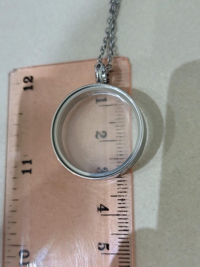 May include: A silver locket pendant with a clear glass centre, hanging from a fine chain. The locket is placed over a ruler, with measurements visible in inches. The background is a neutral beige.