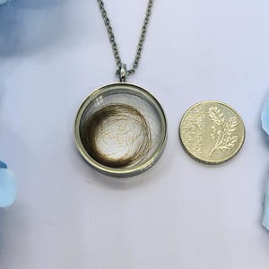May include: A silver pendant necklace featuring a circular glass locket holding a swirl of brown hair. A 5 pence coin is next to the pendant. The chain is visible, and the background is a light blue.