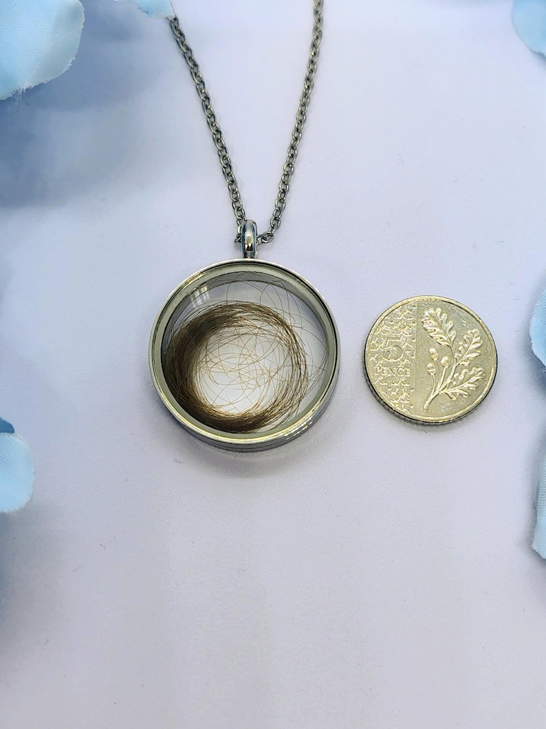 May include: A silver pendant necklace featuring a circular glass locket holding a swirl of brown hair. A 5 pence coin is next to the pendant. The chain is visible, and the background is a light blue.