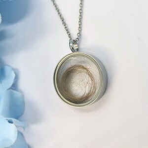 May include: A silver pendant necklace featuring a circular glass locket. The locket holds a small amount of light brown hair. The necklace is set against a white backdrop with a blue floral accent.