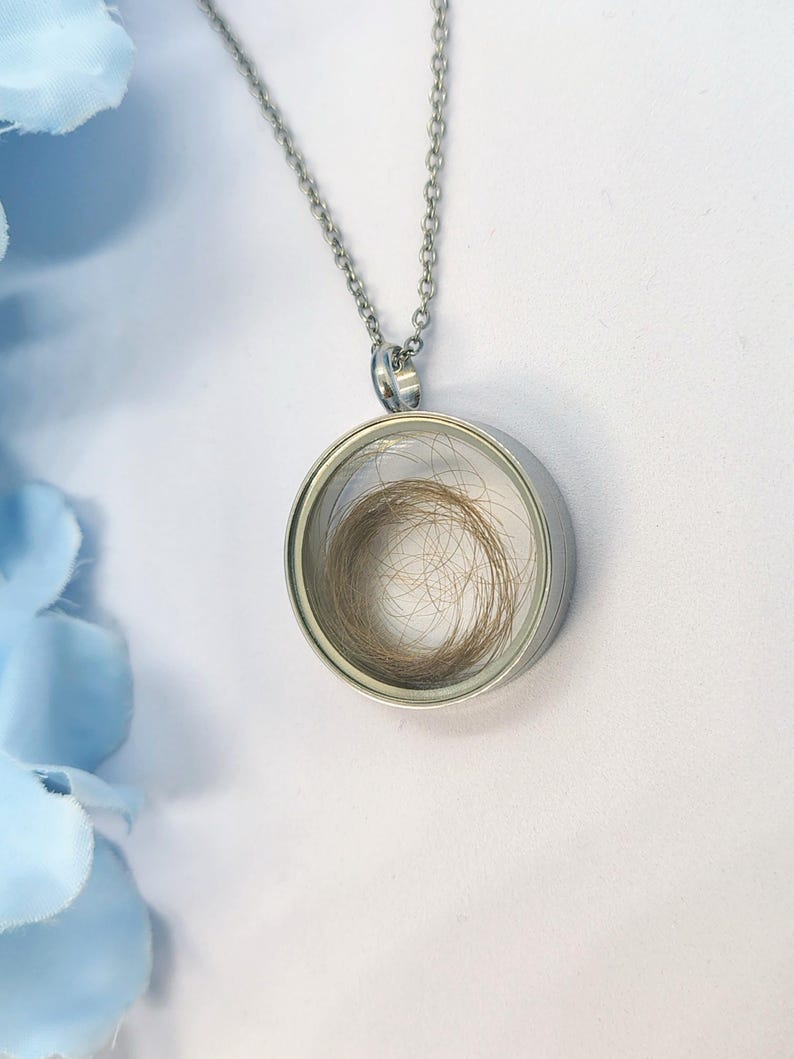 May include: A silver pendant necklace featuring a circular glass locket. The locket holds a small amount of light brown hair. The necklace is set against a white backdrop with a blue floral accent.