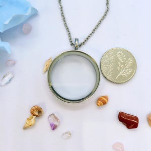 May include: A silver-coloured, round locket necklace with a clear glass centre, displayed with small seashells, gemstones, and a coin. The necklace hangs on a delicate chain. The background is white with light blue accents.