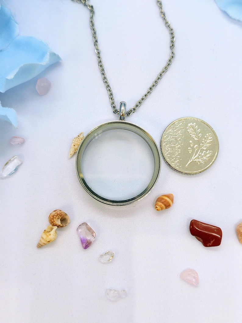 May include: A silver-coloured, round locket necklace with a clear glass centre, displayed with small seashells, gemstones, and a coin. The necklace hangs on a delicate chain. The background is white with light blue accents.