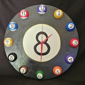 May include: A round clock with a dark gray face, featuring billiard balls as hour markers. The center displays the number 8 in black, with red clock hands. The clock's design is reminiscent of a pool table.