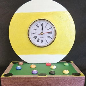 May include: A novelty clock with a round face featuring Roman numerals and a gold-colored bezel. The clock is set into a yellow and white painted wooden billiard ball design. The base is a miniature pool table with colorful billiard balls.