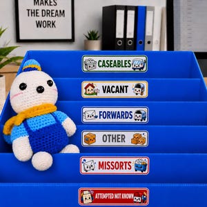 May include: A blue, multi-tiered organiser with labels for mail sorting. A crocheted doll with a blue outfit and yellow scarf sits in the organiser. Labels include "Caseables", "Vacant", "Forwards", "Other", "Missorts", and "Attempted Not Known". A framed sign reads "Makes the Dream Work".