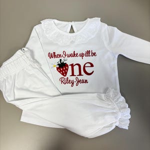 May include: White long-sleeved pajama set with a lace-trimmed collar and cuffs. The shirt features the text "When I wake up I'll be one" with a strawberry graphic and the name "Riley-Jean" in red script. The pants are white.