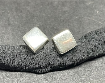May include: A pair of square, silver-colored stud earrings. Each earring has a slightly rounded, square shape. The earrings are displayed on a black, textured surface.