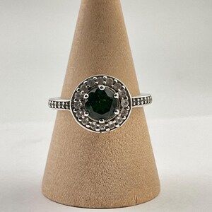 May include: A silver ring with a large, round, dark green gemstone at the center, surrounded by a halo of smaller, clear stones. The band features a textured design. The ring is displayed on a brown, cone-shaped stand.