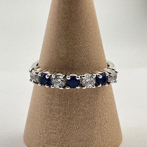 Puede incluir: Un anillo plateado con piedras preciosas azules y claras alternadas. El anillo se exhibe sobre un soporte cónico marrón. Las piedras son redondas y están espaciadas uniformemente.