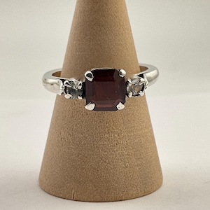 Puede incluir: Anillo de plata con una gema roja oscura cuadrada grande y dos gemas claras más pequeñas. El anillo se exhibe sobre un soporte cónico marrón. La banda del anillo es lisa y pulida, sobre un fondo neutro.