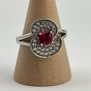 May include: A silver ring with a square, deep red gemstone at the center, surrounded by smaller, clear stones. The ring's design features two circular clusters of clear stones on either side of the central gem.