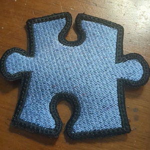 May include: A blue puzzle piece patch with a black border. The patch is made of a woven fabric and has a textured appearance. The puzzle piece shape is a classic symbol of connection and problem-solving.