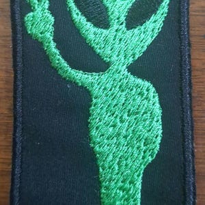 Op de afbeelding: Groen alien geborduurd patch met zwarte ogen en een vredesteken. De alien staat op een zwarte achtergrond.