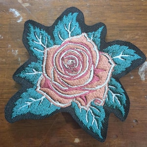 May include: A pink and white embroidered rose with teal green leaves on a black fabric background. The rose is a traditional style with a center swirl and multiple layers of petals.
