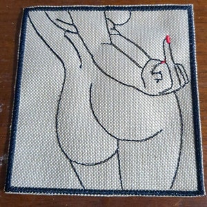 Op de afbeelding: Een beige stoffen patch met zwarte borduurwerk dat een naakt figuur toont dat de middelvinger opsteekt. De vingernagel van de figuur is rood.