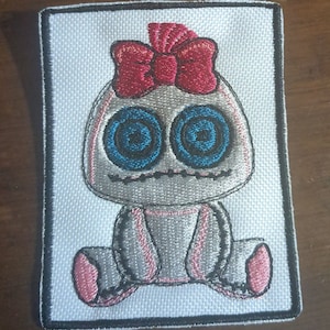 May include: Embroidered patch depicting a seated figure with a skull-like face. It has large blue eyes, a red bow, and a black outline. The patch is rectangular with a black border.