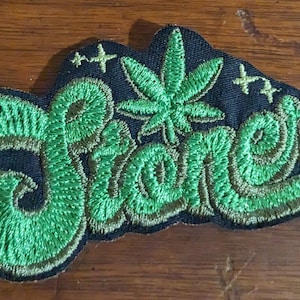 May include: Green embroidered patch with the word "Stoner" in a stylized font. A green marijuana leaf is in the center of the patch.