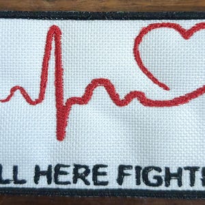 Könnte beinhalten: Weißer gestickter Aufnäher mit einem roten Herzen, einer roten Herzschlaglinie und einem blauen Semikolon. Der Text "STILL HERE FIGHTING" ist unten in Schwarz gestickt.