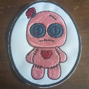 May include: A pink embroidered patch of a voodoo doll with blue eyes, a red heart, and stitched details. The doll is surrounded by a white oval with a black outline.