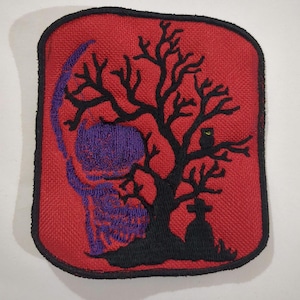 Pode incluir: Um remendo bordado vermelho com uma silhueta preta de uma árvore, uma caveira, uma lápide e uma coruja. A caveira é roxa e a coruja tem olhos amarelos.