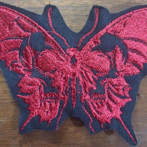 May include: Embroidered patch featuring a red butterfly design on a black background. The butterfly has detailed wing patterns and a skull-like face. The patch is likely for clothing or accessories.