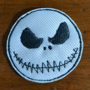 May include: White fabric patch with black embroidered details of a grinning face with a stitched mouth and two large eyes.