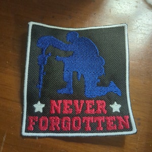 May include: A black embroidered patch with a blue silhouette of a soldier kneeling and holding a rifle. The text "NEVER FORGOTTEN" is embroidered in red below the soldier.