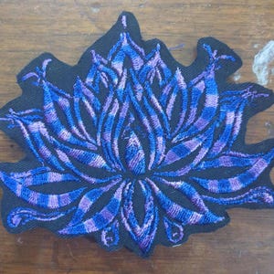 May include: A purple and blue embroidered lotus flower patch. The patch is on a black fabric background.