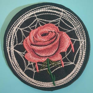 May include: A pink rose embroidered on a black background with a white spiderweb design. The rose is in the center of the circle and has a green stem.