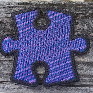 Puede incluir: Una pieza de rompecabezas de color morado y azul con un contorno negro. La pieza está hecha de una tela suave y texturizada.